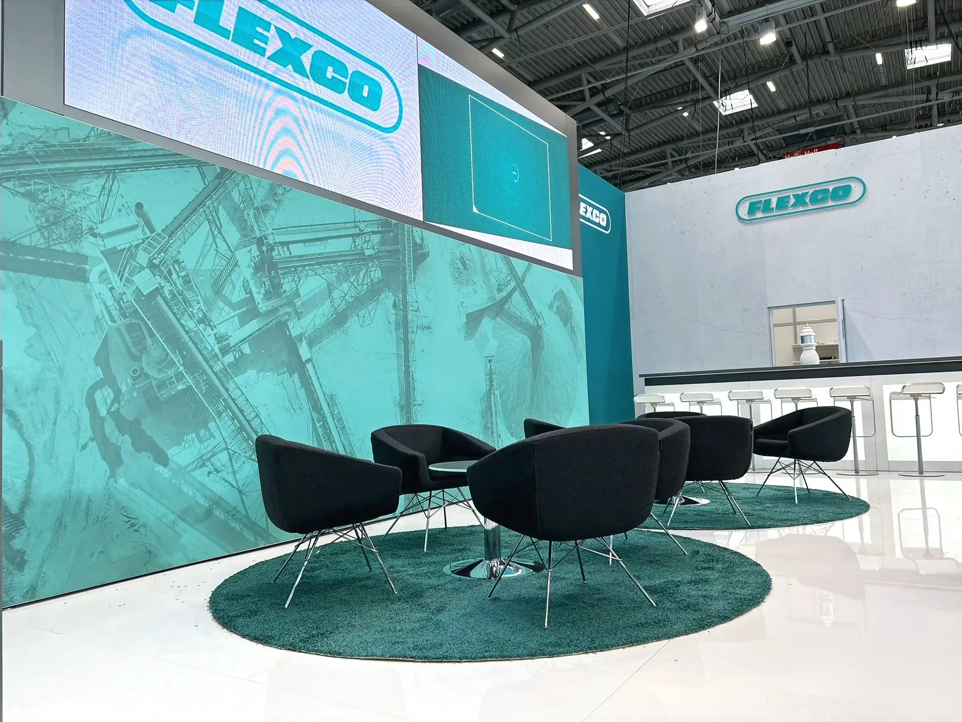 "Moderner Messestand der Firma FLEXCO mit weiß-türkisem Design, großem Logo, Multimedia-Bildschirm und ausgestellten technischen Produkten. Präsentationsflächen mit klaren Linien zeigen Förderbandlösungen und Komponenten. Hängende Deckenbanner und prominente Kontaktdaten runden das professionelle Erscheinungsbild ab."