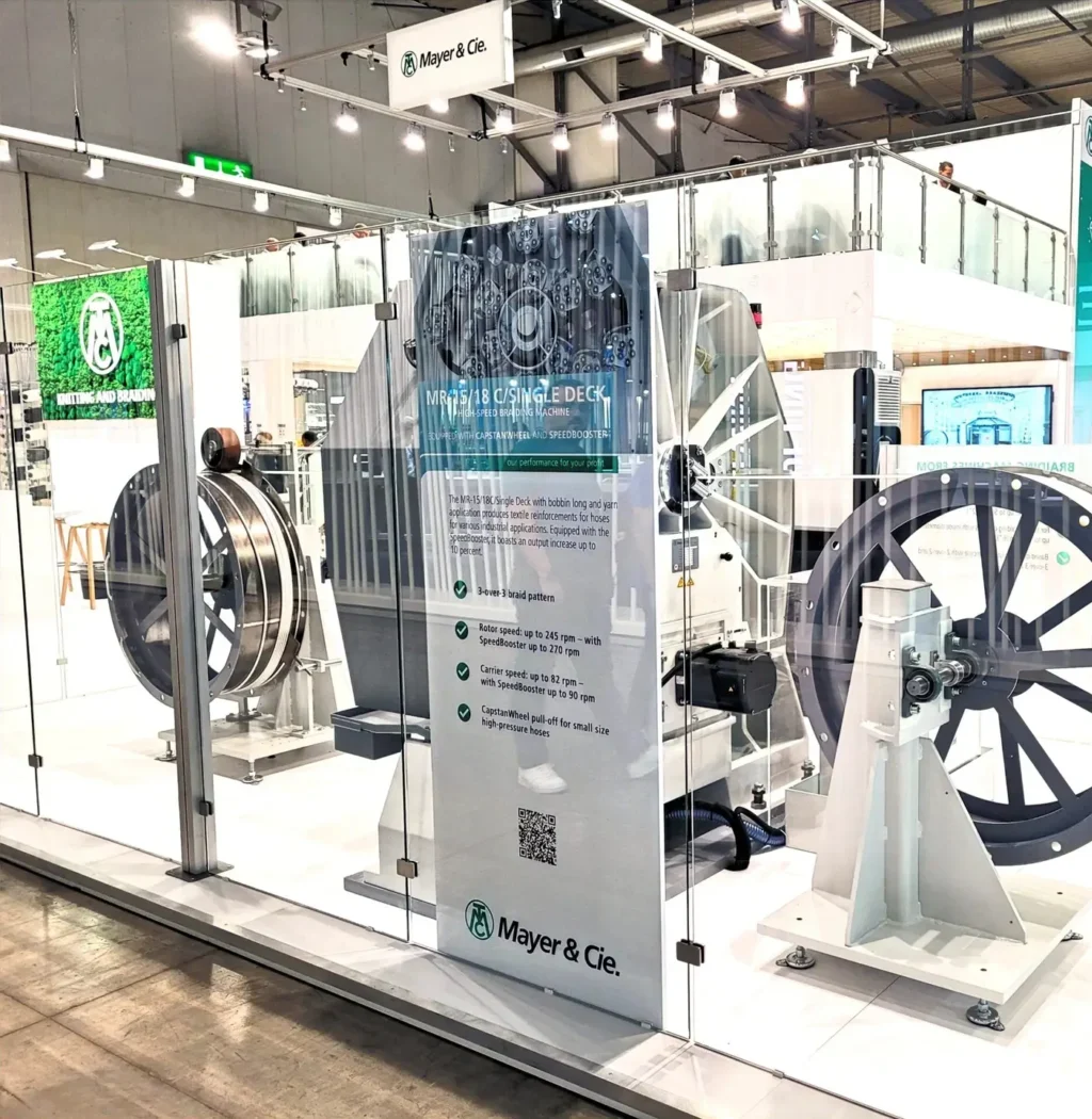 Großer Doppelstock-Messestand mit einer Fläche von 400 qm und einer zweiten Ebene von 100 qm. Offenes, modernes Design mit Glaswänden, großzügigen Ausstellungsflächen und markantem Branding der Firma Mayer & Cie. Der Stand zeigt industrielle Maschinen, informative Displays und Sitzbereiche auf beiden Ebenen. Helle Beleuchtung und klare Strukturen laden Besucher ein.